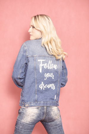 one young woman, looking sideways, 20-29 years old, long blond hair. Shot in studio on pink background. Wearing jeans jacket with sign "follow your dreams" on her back, (rear view).の写真素材