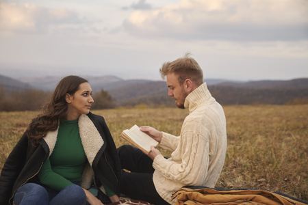 two young people, 20-29 years old, couple or friends, sitting on mountain field outdoors, relaxing, man reading a book, woman listening.の写真素材