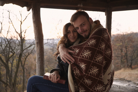 two people, young couple, hugging under a blanket, in rural area. In national park "Fruska Gora", Serbia, sitting under a wooden, summer house, outdoors in Autumn.の写真素材