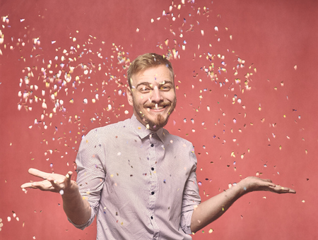 one young smiling man, 20-29 years, confetti falling down, while his arms are outs outstretched. he is looking friendly and welcoming. shot in studio on pink background.の写真素材
