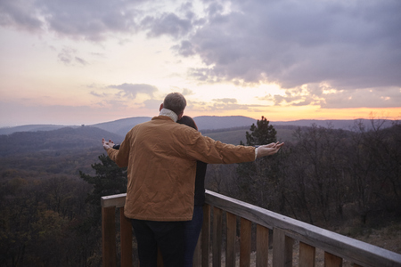 two young people couple, man standing behind woman, holding hands together. rear view, back. Like in a popular movie, on a viewpoint looking at national park "Fruska Gora", Serbia.の写真素材