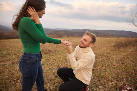 Young man kneeling proposing to young woman, his fiancee. Romantic couple age range of 20-29 years...