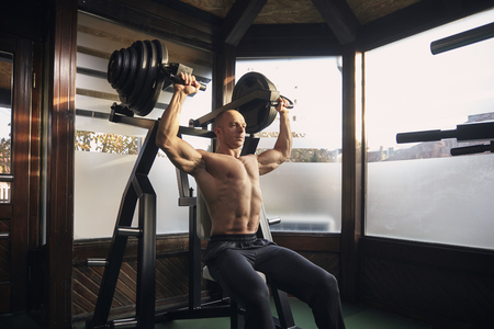 one man, 30-39 years, bodybuilder exercising overhead plate press machine. At home (with many windows) in his own gym with professional fitness equipment.の写真素材