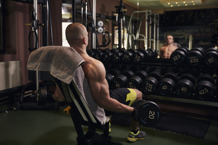 One muscular man bodybuilder, rear view (from behind), sitting on bench, training with dumbbells, indoors in home gym in a room. Whole row of dumbbells in front of large glass.の写真素材