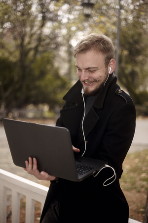 one young man, 20-29 years, smiling and happy, while he is holding laptop in one hand outside in public park, communicating over Internet, video call or chat, could be with family of friend.の写真素材