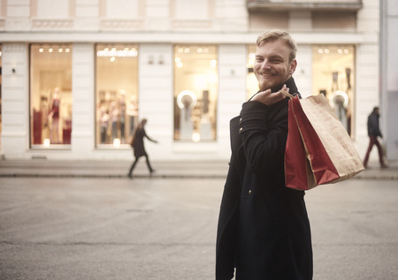one young smiling and happy man, 20-29 years old, handsome and stylish, looking to camera while holding two shopping bags on his back. Pedestrians walking in background, store fronts windows included.の写真素材