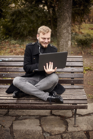 one young relaxed and smiling man, sitting casually on bench with crossed legs in public park, using laptop holding it with one hand. Formal wear or smart casual. Full length shot.の写真素材