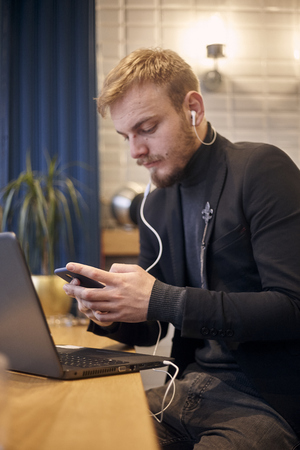 foreground focus on hand and smart phone, man is out of focus. one young man, sitting indoors in coffee shop and using his smart-phone, with laptop nearby, earbuds in ear.の写真素材