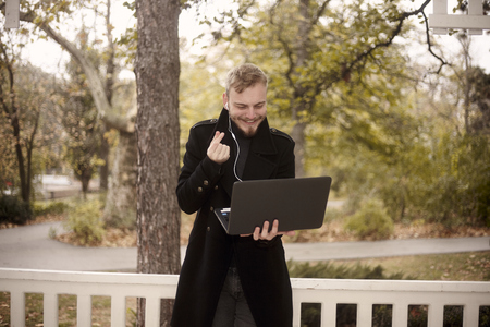 one young man, smiling and happy, holding laptop in one hand outside in park, communicating over Internet, video chat, could be with family of friend, gesturing with hand (Korean heart )の写真素材