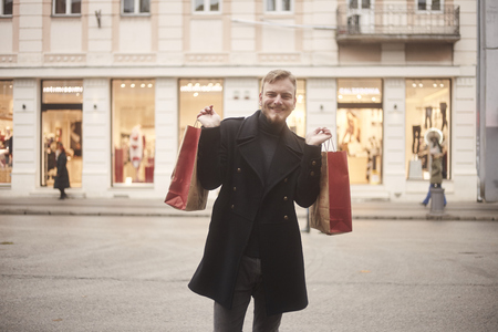 one young smiling and happy man, 20-29 years old, on a pedestrian street, looking to camera, while holding two shopping bags with arms outstretched. Store front windows in distance with pedestrians.の写真素材