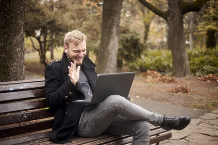 one young laughing man, sitting on bench in public park,  using laptop, talking over Internet, video chat or call, could be with family, friend. gesturing "Hello!" with waving hand.の写真素材