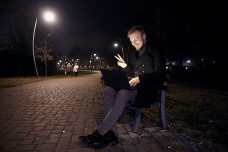 dark night, one young man, 20-29 years, sitting on bench alone, using his laptop in public park in Autumn. gesturing Hello or Hi, with two fingers as a greeting, over video conference call or chat.の写真素材