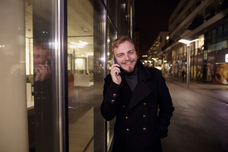 night time, one young smiling and happy man, 20-29 years, talking over his phone, standing in Autumn coat on street, in front of business offices windows outdoors, looking to camera.の写真素材
