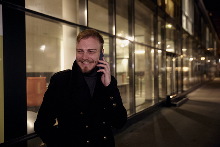 night time, one young smiling and happy man, 20-29 years, talking over his phone, standing in Autumn coat on street, in front of business offices windows outdoors.の写真素材