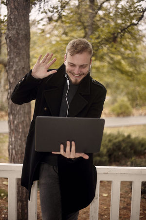 one young smiling and happy man holding laptop in hand, greeting someone (gesturing with one raised hand) over video conference call, could be communicating with family or as businessman, entrepreneurの写真素材