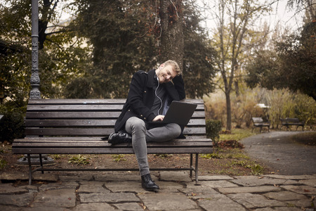 one young smiling and happy man, sitting on bench in public park,  using laptop, looking to computer. Formal wear or smart casual. Full length shot.の写真素材
