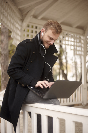 one happy young man, 20-29 years old, sitting on fence outdoors in park, while using his laptop (autonomously without power source) and typing on keyboard, with in-ear earbuds.の写真素材
