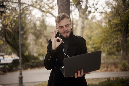 one young man, holding laptop in one hand outside in park, communicating over Internet, video chat or call, could be with family, friend or business, gesturing with hand and two fingers it's perfect!の写真素材