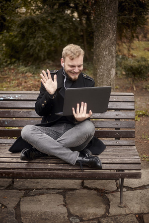 one young relaxed and smiling man, sitting casually on bench with crossed legs in public park, using laptop with video call or video chat, waving hand "Hello" or "Hi!" gesture. Full length shot.の写真素材