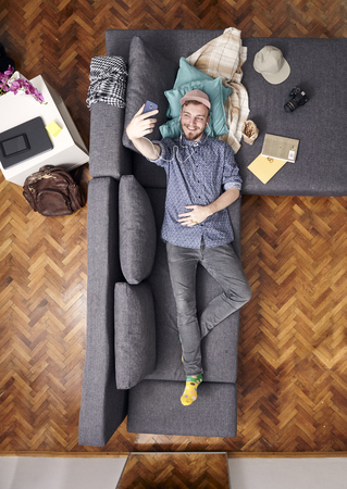 one young smiling man, talking selfie with his smart-phone, laying in sofa. view from above of his whole room, full lenght shot.の写真素材