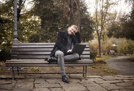 one young relaxed man, sitting on bench in public park, using laptop, looking to camera. Formal wear or smart casual. Full length shot.の写真素材