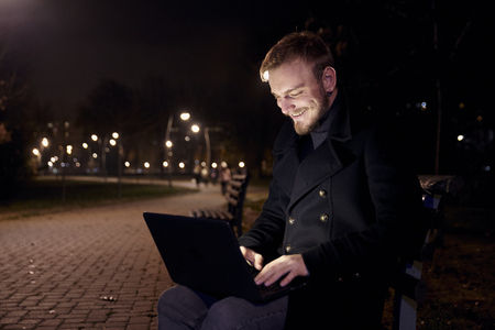 dark night, one smiling young man, 20-29 years, sitting on bench alone, using his laptop in public park in Autumn.の写真素材