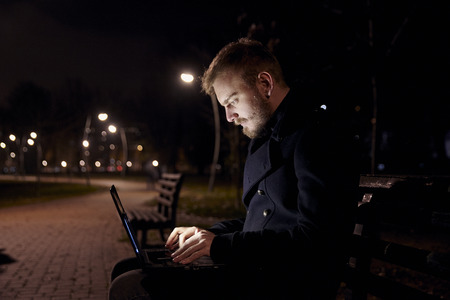 dark night, one young man, 20-29 years, sitting on bench alone, using his laptop typing on keyboard, in public park in Autumn.の写真素材