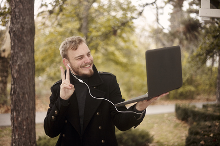 one young man, smiling and happy, holding laptop in one hand outside in park, communicating over Internet, video chat, could be with family of friend, gesturing with hand (two fingers as peace sign)の写真素材