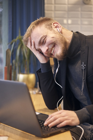 one young candidly smiling man, sitting indoors in coffee shop and using his laptop, earbuds in ear.の写真素材