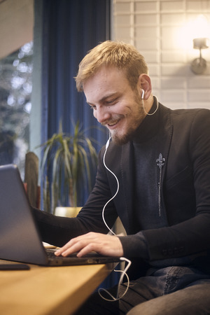 one young smiling man, sitting indoors in coffee shop and using his laptop, earbuds in ear.の写真素材