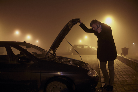 One young man, talking over phone, while holding car hood and looking at engine. It's dark and nighttime, with mist and fog, rural area.の写真素材
