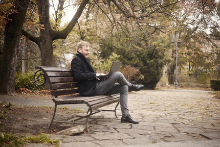one young man, sitting on bench in public park,  using laptop, talking over Internet, video chat or call, could be with family, friend or business.の写真素材