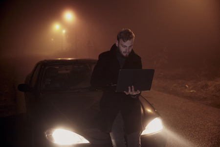 one young man using his laptop computer, on a car hood. It's dark and night time, in rural area.の写真素材