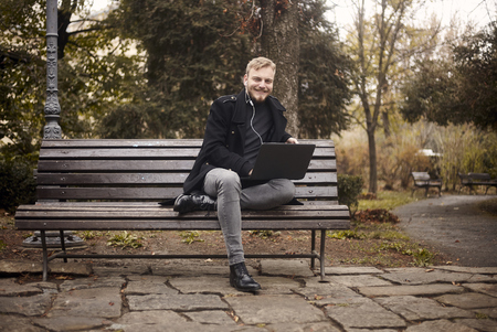one young smiling and happy man, sitting on bench in public park,  using laptop, looking to camera. Formal wear or smart casual. Full length shot.の写真素材