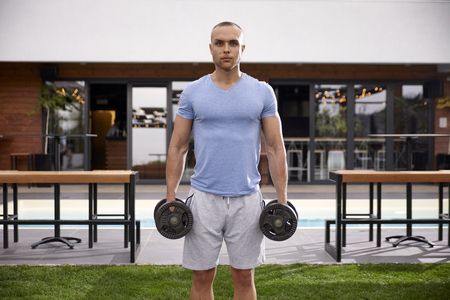 One young man, 20-29 year old, standing in front of the camera while holding two weights outside.Front view.の写真素材