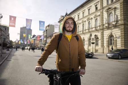 one young man, 20-29 years old, wearing jacket and hoodie, pushing his bicycle in city, pedestrian area (urban area). Looking to camera.の写真素材
