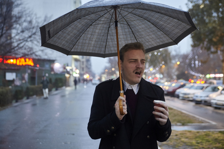 one young businessman, 20-29 years old, yawning while holding an umbrella and a cup, while going to a work.の写真素材