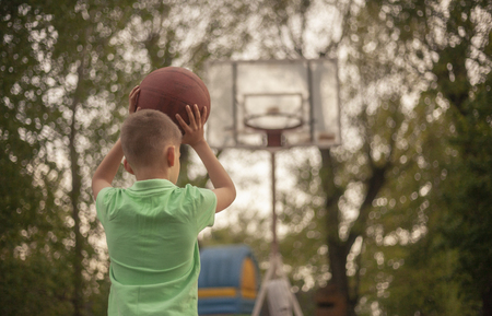One nine years old boy, rear view from behind, holding a ball in his hands, raising it up to shoot. On a Spring day, at a basketball court outdoors.の写真素材