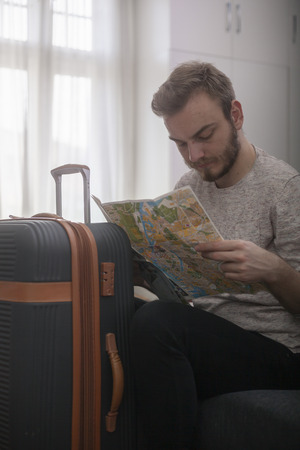 one young man - traveler looking closely at city map, while sitting near a suitcase, indoors.の写真素材
