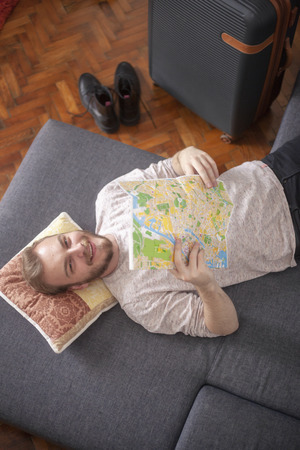 One young man - traveler holding a city map, while looking at camera, while resting indoors on a bed. Suitcase nearby.の写真素材