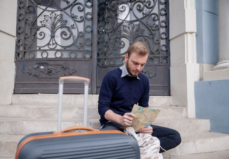 One young lost traveler or tourist resting on a steps with a suitcase, while looking at a street map...