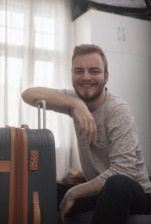 one young man - traveler sitting in a room. Looking at a camera, smiling, while resting his arm on a suitcase.の写真素材