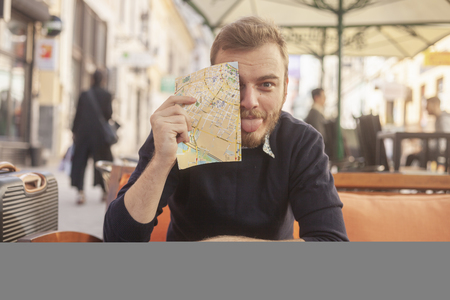 one young man - happy traveler or tourist, making a face, with a tongue out. Looking silly to a camera. Sitting in a cafe, outdoors on a street.の写真素材