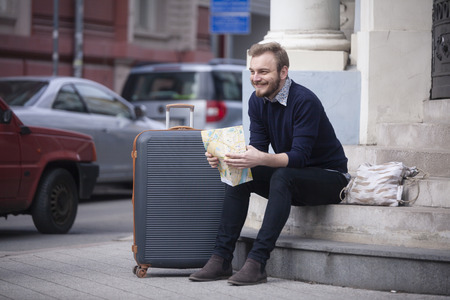 one young happy man, tourist or traveler, sitting on a steps on a street, with a city map in his hands, with a suitcase nearby.の写真素材