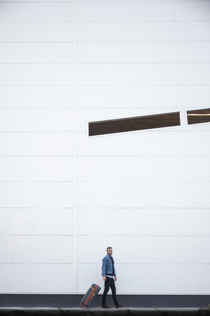 one young man walking with a suitcase. Behind him is a building exterior large modern wall. In comparison man is small.の写真素材