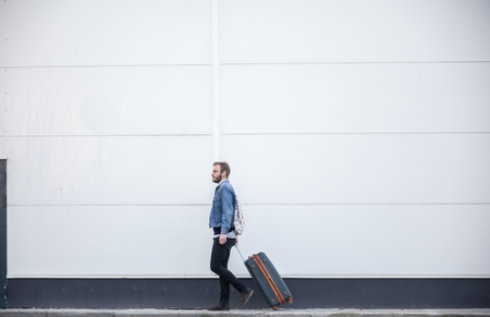 one young man - traveler or tourist, walking with a suitcase. building exterior white wall behind, minimalistic style.の写真素材