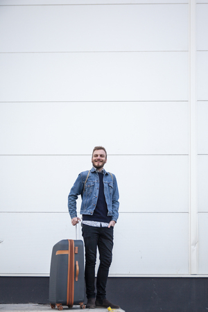 one young man, 20-29 years, traveler outdoors happy, with a suitcase. Behind is a white building exterior wall minimalist white.の写真素材