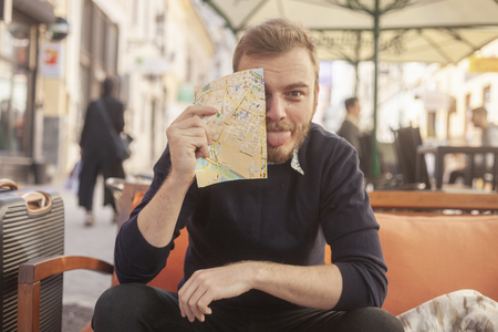 one young man - happy traveler or tourist, making a face, with a tongue out. Looking silly to a camera. Sitting in a cafe, outdoors on a street.の写真素材