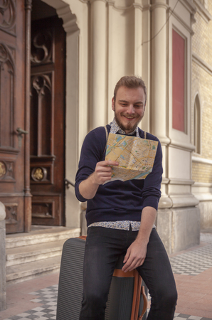 one young man - traveler or tourist, looking at city map while sitting on a suitcase. relaxing in a old European architecture style, outdoors.の写真素材