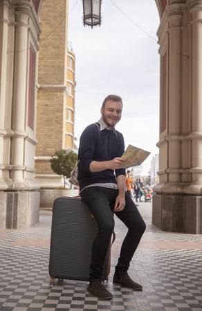 one young man - traveler or tourist, looking at a camera, while holding city map and sitting on a suitcase. relaxing in a old European architecture style, outdoors.の写真素材